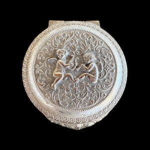 May include: Silver-toned, round compact with a hinged lid. The lid features a detailed relief design of two cherubs with bows and arrows, surrounded by ornate floral patterns. The compact has a decorative border.