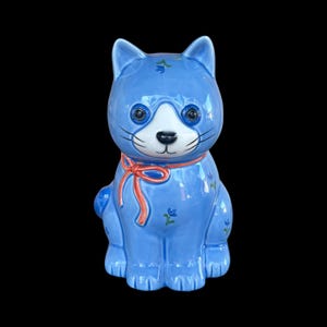 May include: A ceramic cat figurine in a sitting position. The cat is light blue with white and black facial features, including large eyes and whiskers. It has a red bow around its neck and small floral accents.