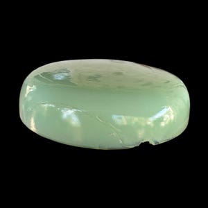 May include: A pale green, oval object with a shiny, smooth surface. The object has a slightly uneven shape with a small chip on one side. The background is plain black.