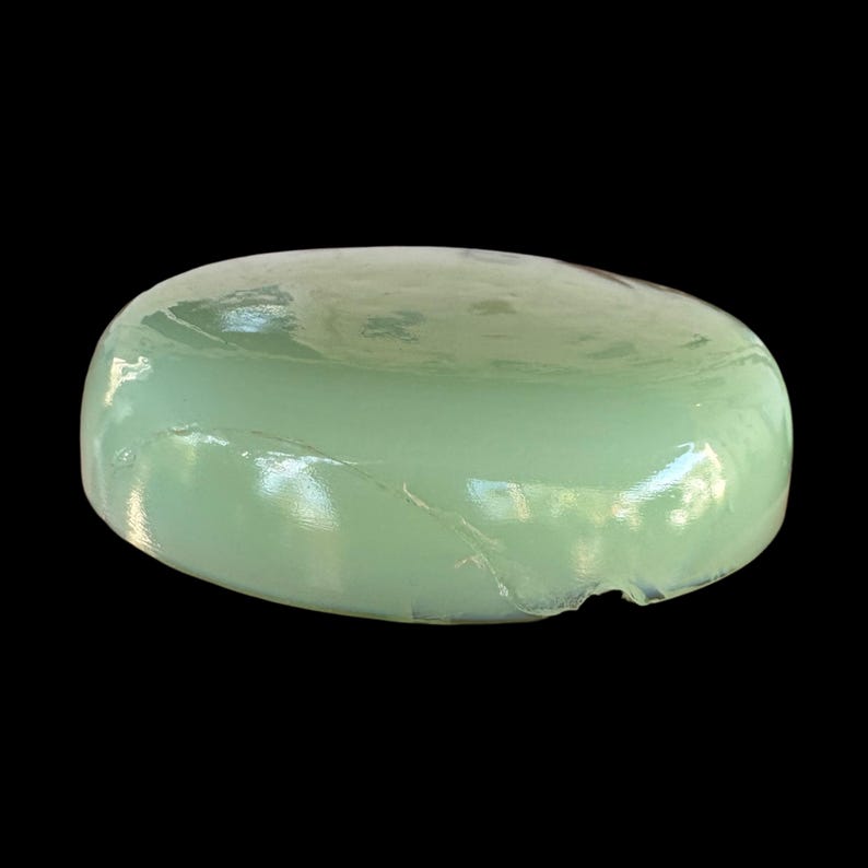 May include: A pale green, oval object with a shiny, smooth surface. The object has a slightly uneven shape with a small chip on one side. The background is plain black.