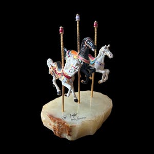 May include: A carousel sculpture with three horses in white and black, adorned with colourful saddles and gold poles. The sculpture is mounted on a light-coloured stone base. The text "103/5000" is visible on the base.