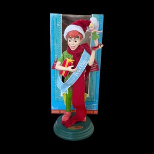 May include: A Peter Pan figurine wearing a Santa hat, green shirt, red scarf, and green pants, holding a wrapped gift. A banner reads "Magical Peter Pan". Tinkerbell is on his right hand. The figurine is on a green base.