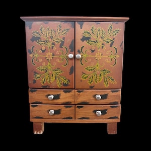 May include: A small wooden cabinet with two doors and four drawers. The cabinet is brown with black distressed accents and a floral design in yellow and green. Silver knobs adorn the doors and drawers. The cabinet sits on short legs.