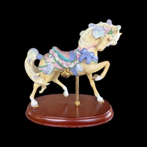 Könnte beinhalten: Eine cremefarbene Karussellpferde-Figur, verziert mit pastellfarbenen rosa, blauen und grünen Akzenten. Das Pferd hat einen blumigen Sattel, eine blaue Schleife und eine goldfarbene Stange, montiert auf einem runden, dunkelbraunen Holzsockel.