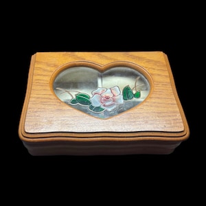 May include: Wooden jewelry box with a heart-shaped glass window. The window features a painted rose with pink petals and green leaves. The box is rectangular with a hinged lid and a light brown finish.