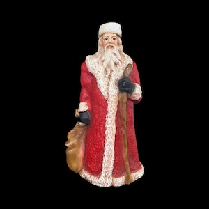 May include: A figurine of Santa Claus, dressed in a red coat with white fur trim, a white hat, and black gloves. He carries a wooden staff and a tan sack. The figure has a long white beard and is set against a black background.