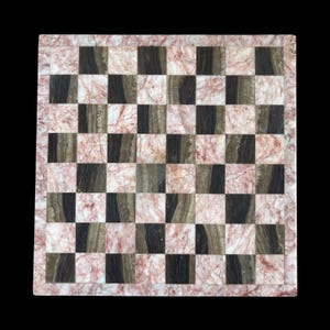 May include: A square chessboard featuring a checkerboard pattern of alternating dark brown and light pink marble tiles. The board is framed by a border of the same pink marble, creating a smooth, polished surface with natural stone veining.