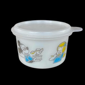 May include: A white plastic bowl with a lid, featuring cartoon illustrations of Mickey Mouse and a girl. The bowl has a handle and is decorated with blue, yellow, and black accents. The bowl is likely vintage.