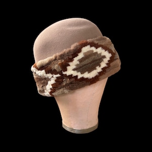 May include: A tan cloche hat with a decorative band. The band features a geometric pattern in shades of brown and cream. The hat is displayed on a mannequin head against a black background.
