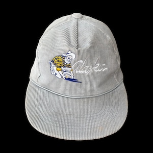 Op de afbeelding: Een lichtgrijze baseballpet van corduroy met een geborduurde afbeelding van een persoon met een baard in een geel en blauw geruit overhemd die naar goud zoekt. Het woord Alaska is in witte cursieve letters rechts van de figuur geborduurd.