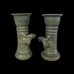 May include: Two green ceramic candle holders. Each holder features a frog design on the side. The candle holders have a flared top and a round base. The frog is sitting on a leaf. The candle holders are a muted green color.