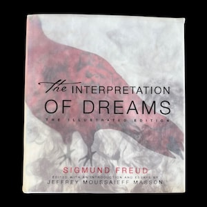 May include: A hardcover book titled The Interpretation of Dreams: The Illustrated Edition by Sigmund Freud, edited by Jeffrey Moussaieff Masson. The cover features a large, dark red bird silhouette against a white, smoky, and cloudy background.