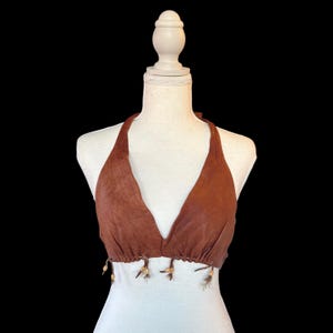 May include: A brown halter top made of a soft, textured material. The top features a V-neck design and is adorned with small, decorative beads and feathers along the bottom hem. The top is displayed on a white mannequin against a black background.