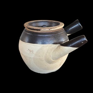 May include: A two-toned ceramic teapot with a dark brown top and spout, and a cream-colored base. The teapot has a round body, a lid, and two spouts extending from the side. The background is black.