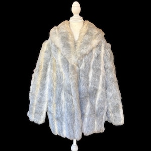 May include: A long-sleeved faux fur coat featuring a vertical striped pattern in shades of light gray and white. The garment has a wide, plush collar and a straight hemline, displayed on a white mannequin against a solid black background.