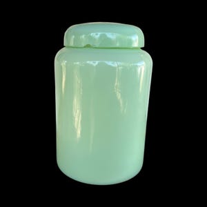 May include: A pale green glass jar with a matching lid. The jar is cylindrical with a rounded base and a lid that fits securely. The glass has a smooth, glossy finish. The jar is set against a black background.