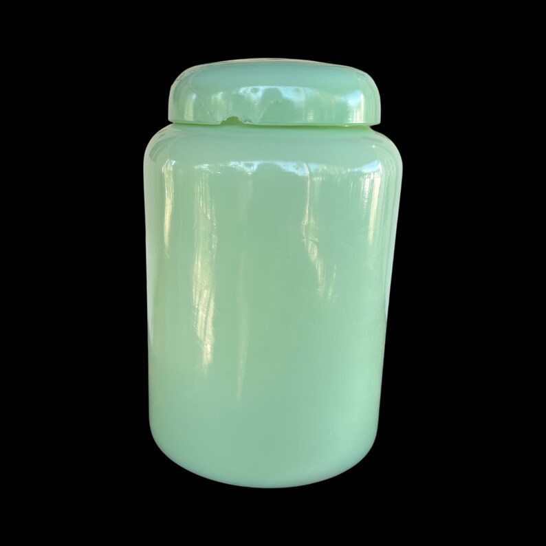 May include: A pale green glass jar with a matching lid. The jar is cylindrical with a rounded base and a lid that fits securely. The glass has a smooth, glossy finish. The jar is set against a black background.