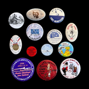 May include: A collection of thirteen assorted circular and oval pinback buttons featuring various graphic designs, illustrations, and text including "Shasta Dixieland Jazz Festival," "Bike Show 2001," "Dogtown Donkey Derby," and "Happy Birthday."