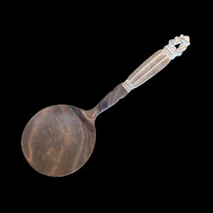 May include: A silver serving spoon with a round bowl and a decorative handle. The handle features a textured design with a detailed finial. The spoon is made of a shiny, reflective metal, likely stainless steel.