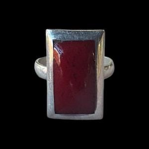 May include: A silver ring with a rectangular, deep red stone. The stone is set within a silver frame, and the band is a simple, rounded design. The ring is set against a black background.