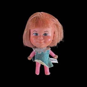 May include: A vintage doll with a smiling face, short orange hair, and blue eyes. The doll wears a teal dress with a silver collar and pink legs. A small tag that says "TAIWAN" is attached to the dress.