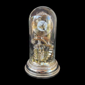 May include: A gold-tone Rhythm quartz anniversary clock encased in a clear glass dome. The clock features a white face with black numerals, floral accents, and a rotating carousel mechanism with metallic figures positioned on a circular base.