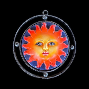 Könnte beinhalten: Ein rundes, handgemaltes Sonnengesicht mit lächelndem Ausdruck, vor blauem Hintergrund. Die Sonne hat orange und gelbe Strahlen und einen roten Mund. Es ist in einem Metallring mit einer Schlaufe zum Aufhängen eingefasst.
