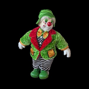 May include: A vintage clown doll in a green jacket with red trim and gold pockets. The doll has a white face, red nose, and green hat. The clown wears a black and white polka dot shirt and striped trousers.
