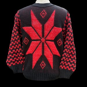 May include: Black knit sweater featuring a large red starburst design on the back. The sleeves have a red and black checkerboard pattern. The sweater has a crew neck and is made of a thick knit material. A festive, winter-themed garment.
