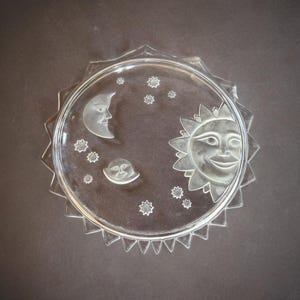 May include: A clear glass plate featuring etched designs of the sun, moon, and stars. The sun has a smiling face, and the moon has a human face. The plate has a decorative, jagged edge.