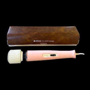 May include: A light pink Hitachi two-speed massager with a white textured head and a power cord, accompanied by a rectangular brown faux-leather storage case featuring the text Hitachi two speed massager printed on the front surface.