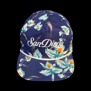 Pode incluir: Um boné de beisebol azul com um padrão floral branco e detalhes em laranja. A parte frontal exibe o texto San Diego em letra cursiva branca. Um cordão trançado branco corre horizontalmente na base da copa, acima da aba do boné.
