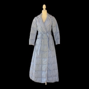 May include: A light blue, floor-length quilted robe featuring a wrap-style front with a matching tie belt. The garment has long sleeves, a notched collar, and white piping trim along the lapels, cuffs, and pockets, creating a structured silhouette.