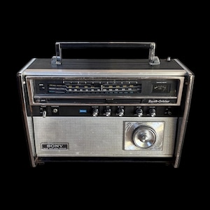 Könnte beinhalten: Ein vintage Sony Earth-Orbiter Multiband-Radioempfänger mit silbernem Metall-Lautsprechergitter, schwarzem Tragegriff und horizontaler Senderskala mit Frequenzanzeigen. Die Frontplatte enthält mehrere Drehregler, einen Netzschalter, eine Kopfhörerbuchse und ein Abstimmungsanzeigeinstrument.