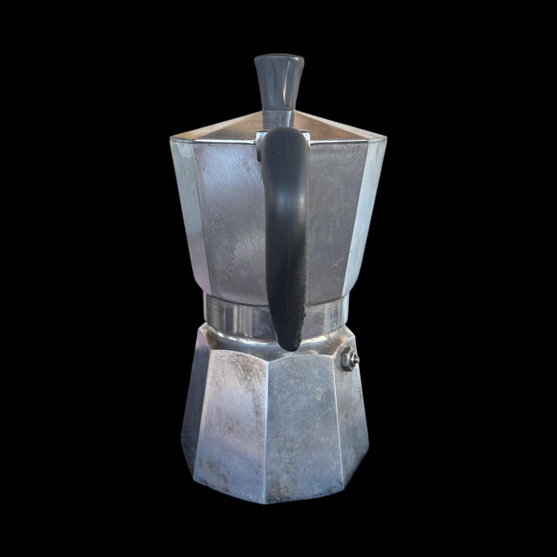 May include: A silver stovetop espresso maker with a black handle and top. The coffee pot has a geometric design and is set against a black background. This Italian-style coffee maker is a kitchen essential.