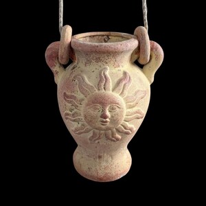 May include: A hanging terracotta-colored ceramic vase featuring a relief of a sun face on the front. The vessel has two side handles with a rope threaded through them, and the surface has a weathered, matte finish with light and dark tones.