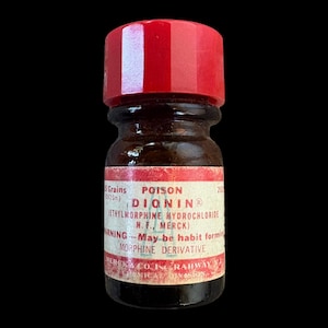 May include: A small brown glass bottle with a red screw-top cap and a white label featuring red text. The label reads POISON, DIONIN, ETHYLMORPHINE HYDROCHLORIDE, MERCK, WARNING - May be habit forming, MORPHINE DERIVATIVE, and MERCK & CO. INC. RAHWAY N.J.