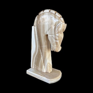 May include: A carved stone sculpture of a horse's head and neck, in shades of beige and brown. The horse is depicted in profile, with detailed features and a flowing mane. The sculpture is mounted on a rectangular base.