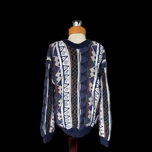 Puede incluir: Un suéter vintage de cuello redondo con cuello y puños azul marino. El suéter presenta un patrón geométrico en tonos de azul, blanco, beige y verde oliva. El diseño incluye rayas verticales, zigzags y secciones a cuadros.