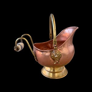 May include: Antique copper and brass coal scuttle with a handle and a decorative lion's head. The scuttle has a copper body, brass accents, and a white and blue ceramic handle. The base is brass and the handle is arched.