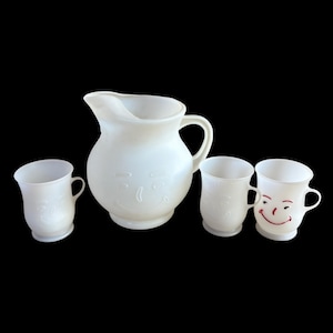 Pode incluir: Uma jarra de vidro leitoso branco e três canecas a combinar. A jarra e duas canecas apresentam rostos sorridentes em relevo. Uma caneca apresenta um rosto sorridente com detalhes pintados a vermelho para os olhos, sobrancelhas e boca.