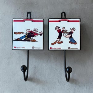 Könnte beinhalten: Zwei schwarze Metall-Wandhaken mit Popeye- und Olive Oyl-Cartoonfiguren. Jeder Haken hat eine quadratische Kachel mit einem Cartoon-Bild und dem Schriftzug "Character Art" oben. Die Haken sind an einer grauen Wand befestigt.