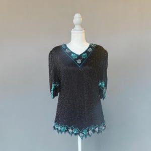May include: Black beaded top with a V-neckline and short sleeves. The top is embellished with teal and silver sequins along the neckline, sleeves, and hemline. The top is displayed on a white mannequin.