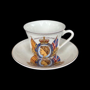 May include: White teacup and saucer set featuring a commemorative design. The cup and saucer are adorned with a portrait, flags, and the text "CORONATION JUNE 2nd 1953". The set has a gold trim.