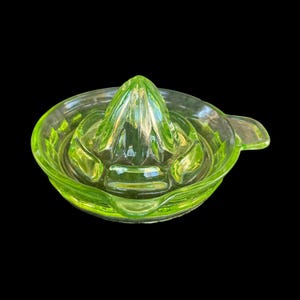May include: A vintage, light green glass citrus juicer. The juicer has a cone-shaped reamer in the center, surrounded by a bowl with a pouring spout. The glass has a ribbed texture, and the overall design is simple and functional.