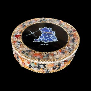 Może przedstawiać: Okrągłe, dekoracyjne pudełko z czarnym wieczkiem z niebieską mapą Brazylii i białym napisem "BRASIL". Pudełko jest wykończone złotym metalem i kolorowymi kamieniami.