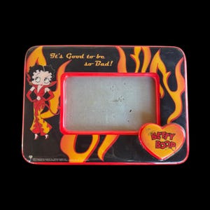 Puede incluir: Marco de fotos rectangular negro con borde rojo y temática de Betty Boop. El marco presenta a Betty Boop con un vestido rojo con llamas amarillas y el texto "It's Good to be so Bad!" y un logotipo en forma de corazón que dice "Betty Boop".