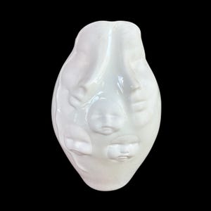 May include: A white ceramic vase with a smooth, glossy finish featuring multiple sculpted human faces integrated into the surface. The vessel has an elongated, rounded shape with several distinct facial features embossed across the front.