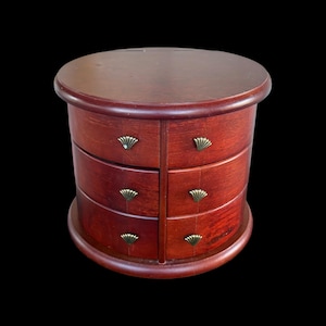 May include: A round, wooden jewelry box with a rich, reddish-brown finish. It features six drawers with decorative, fan-shaped brass pulls. The box has a curved base and a flat, circular top, set against a black background.