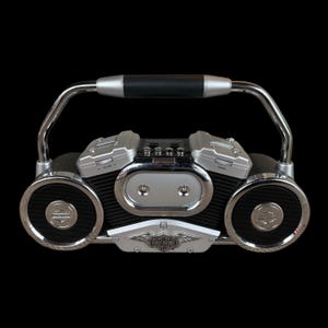May include: A silver and black boombox with a handle. The boombox has two speakers with silver rims and a Harley-Davidson logo. The centre features a silver panel and the Harley-Davidson logo. The handle is silver with a black grip.